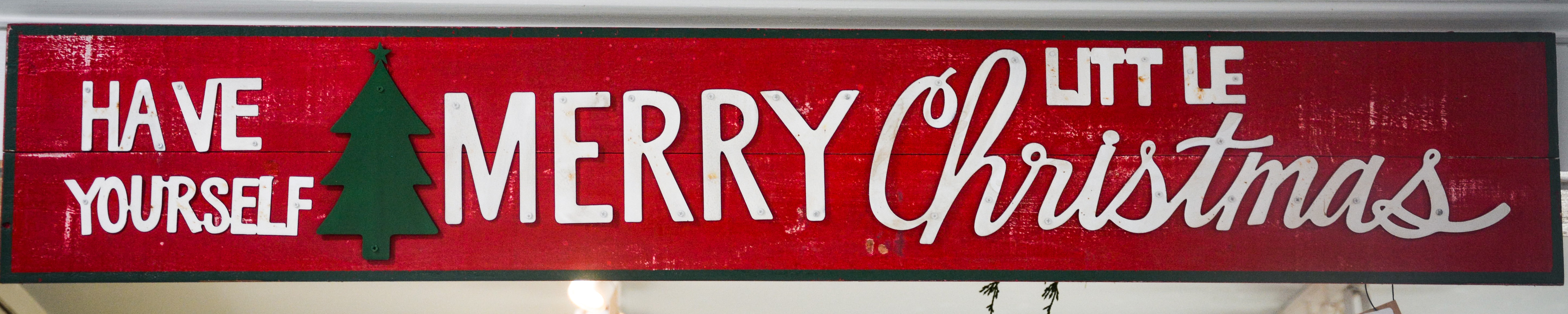 Hand painted red sign with white lettering that reads Have Yourself a Merry Little Christmas, where the letter A is a green Christmas tree.